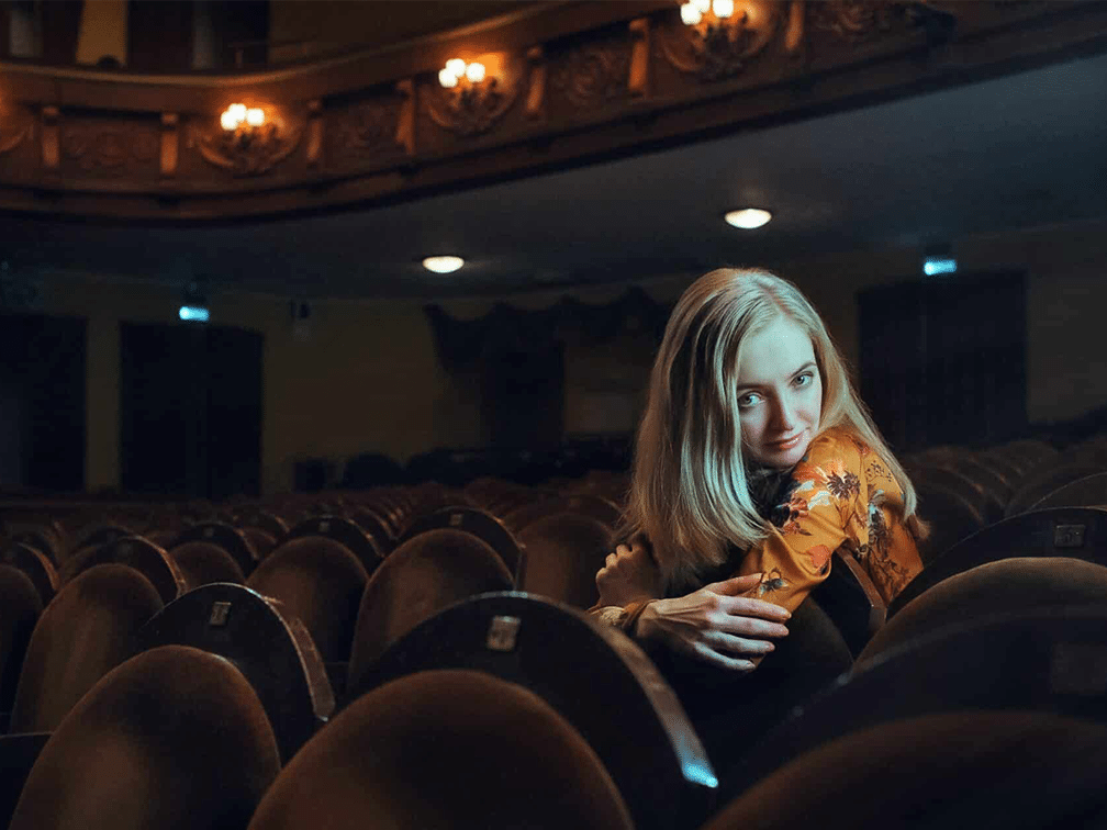 Blonde junge Frau sitzt in einem Theatersaal und lehnt sich über den Sitz in der vorderen Reihe