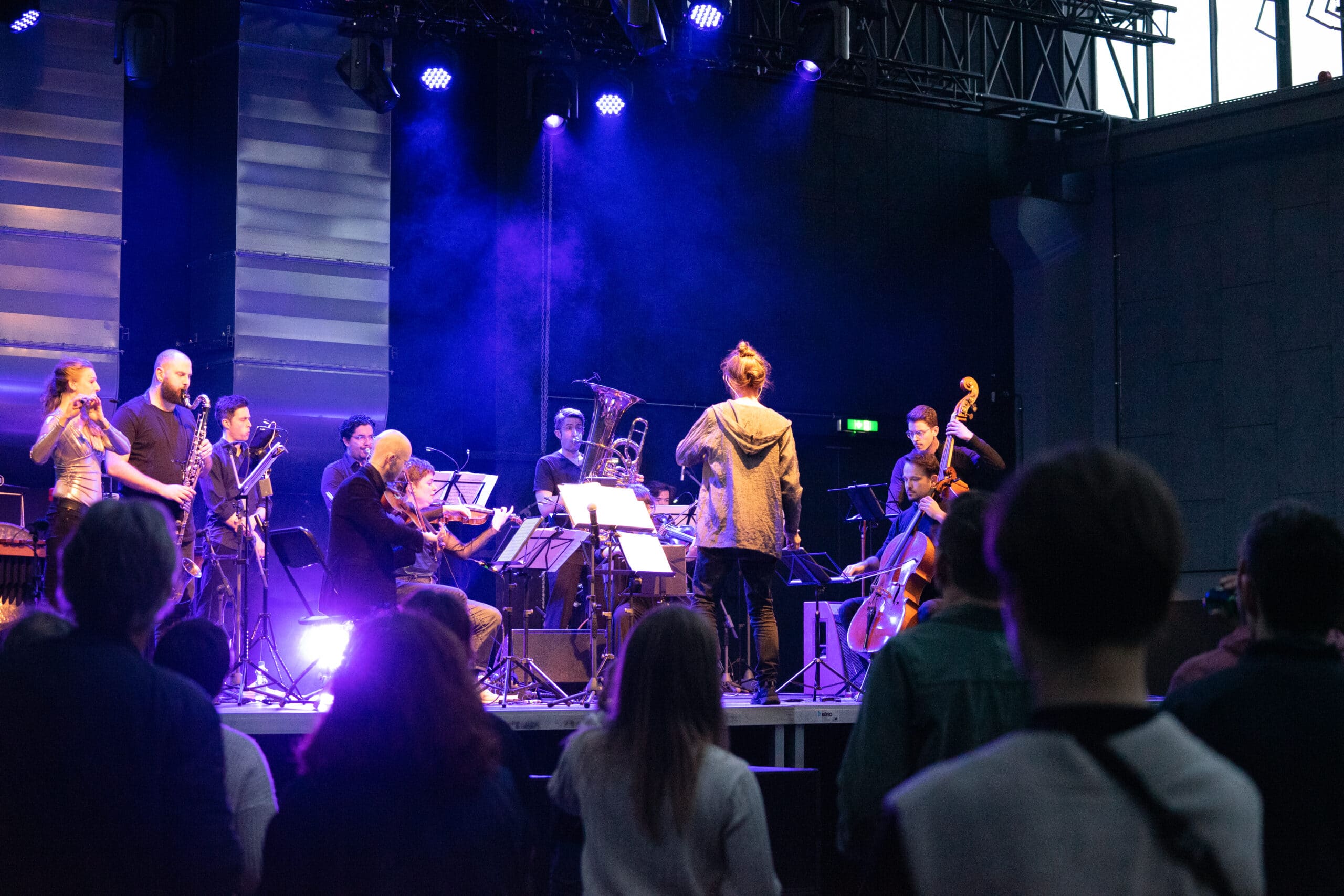 Orchestra in the stairwell plays classical music with club-appropriate sounds in an industrial hall. The audience dances in front of the stage.