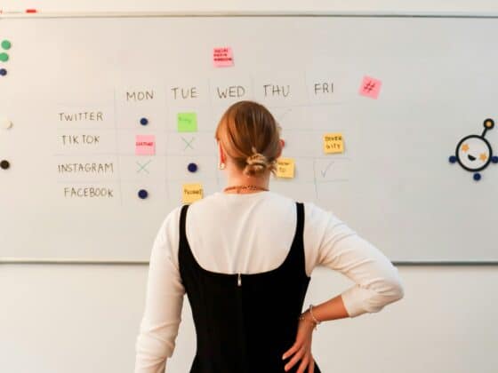 Eine Frau steht nachdenklich vor einem Whiteboard, auf dem ein Wochenplan mit Post-it's und verschiedenen Social-Media-Kanälen zu sehen ist.