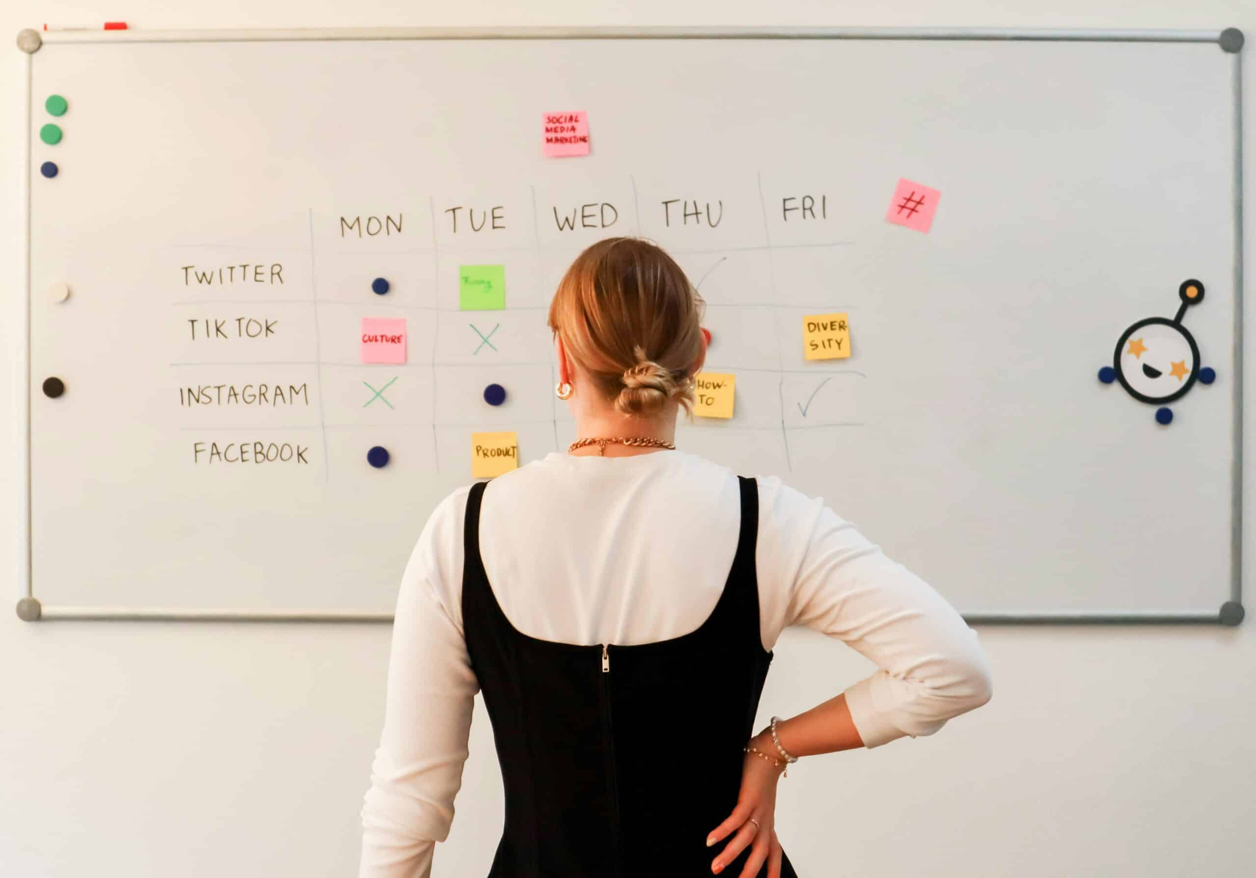 Eine Frau steht nachdenklich vor einem Whiteboard, auf dem ein Wochenplan mit Post-it's und verschiedenen Social-Media-Kanälen zu sehen ist.