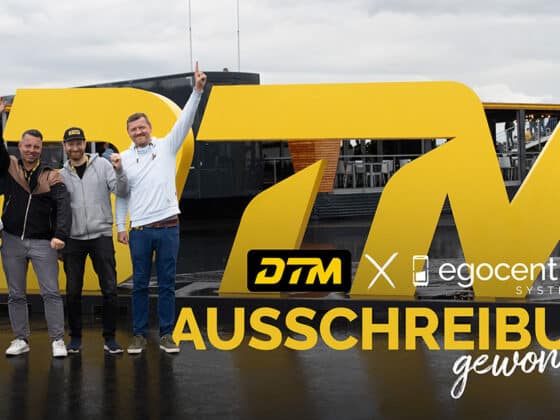 Geschäftsführung und Projektleitung von egocentric Systems stehen vor dem DTM Zeichen an der Rennstrecke und halten feierlich die Arme in die Höhe.