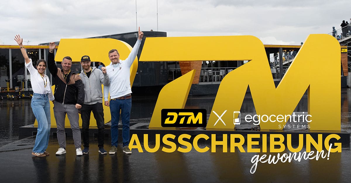 Geschäftsführung und Projektleitung von egocentric Systems stehen vor dem DTM Zeichen an der Rennstrecke und halten feierlich die Arme in die Höhe.